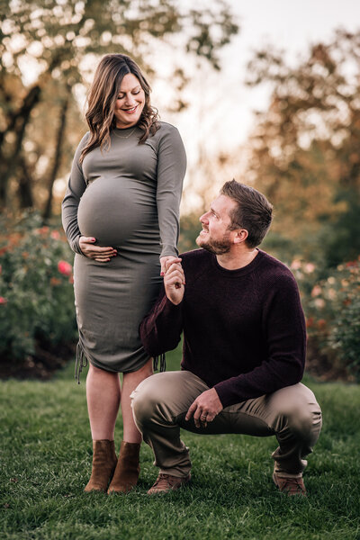 A pregnant woman in a gray dress stands on the grass, holding hands with a man kneeling beside her.