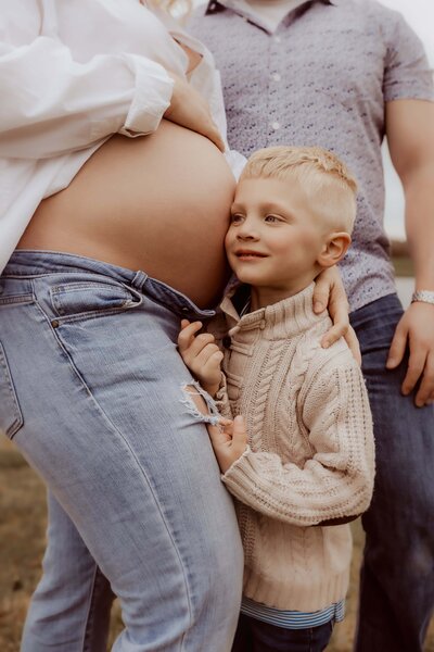 A young child in a sweater touches a pregnant person's belly while standing outdoors. Another adult stands nearby, slightly out of view.