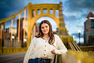 Stunning senior picture in Pittsburgh PA on iconic yellow bridge featuring young woman in white cardigan against downtown skyline.