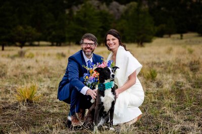A bride and groom crouching down smiling with their dog.