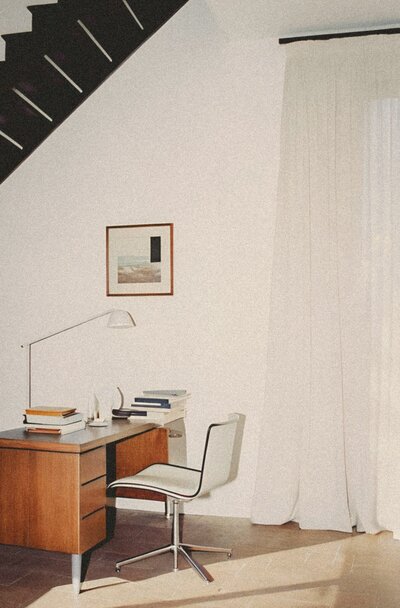 A minimalist white room featuring open black stairs, with a mid-century walnut desk and sleek steel chair beneath. Framed artwork and soft linen curtains add elegance to this thoughtfully curated home office space.