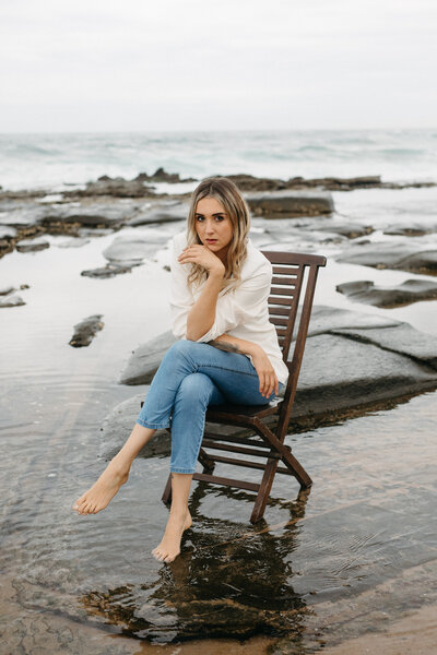 A creative brand photo - the model in jeans and white blazer sits in a chair on the beach. She is looking at the camera and the natural light is soft and bright