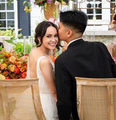 Capture a unique wedding moment with the bride smiling back at the camera while seated beside her groom, who is engaged with friends, at a luxury white tent reception. A perfect blend of joy, elegance, and celebration.
