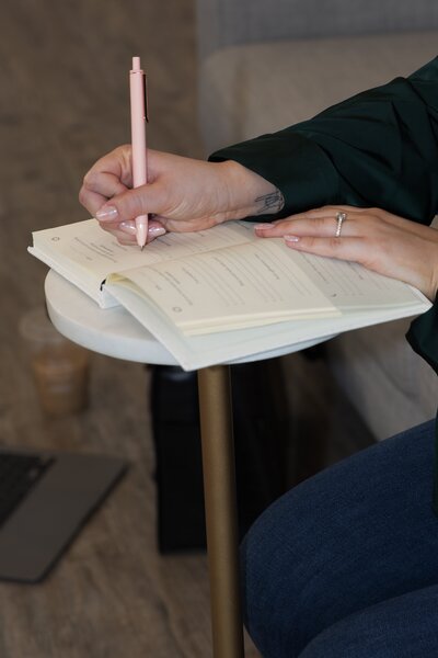 Karleigh Skeans writing in an open notebook. The pages are cream and the pen is light pink. The image is focused on her hands and writing in the notebook.