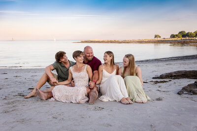 Family of 5 with teenage kids sitting on beach, laughing and smiling at each other.