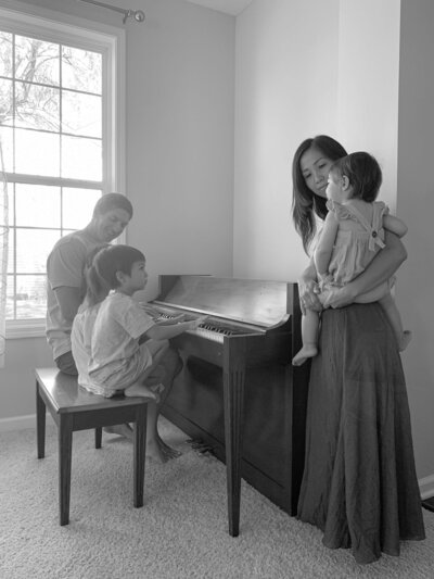 Mai with youngest in her arms, her husband  and two older kids on piano bench playing the piano