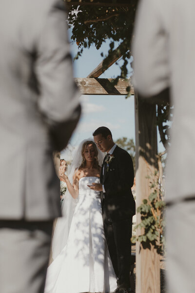 A bride and groom stand together under a wooden arbor during their outdoor wedding ceremony, framed by blurred figures in the foreground as sunlight filters through the scene.