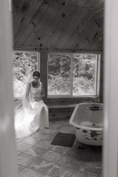 A bride is sitting in a bathroom in a Vermont Airbnb near windows with sunlight shining through. She is wearing a long mesh veil and silky wedding dress.