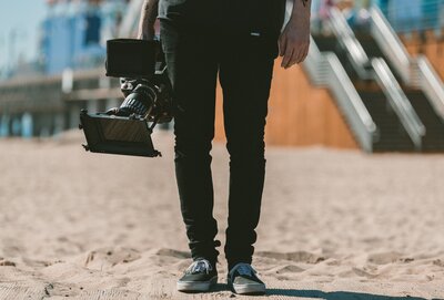 Outdoor video production with videographer carrying professional camera equipment on sandy beach location