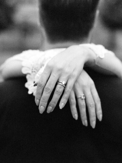 Bride embraces her husband with hands crossed behind him, showing off wedding rings