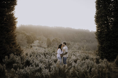 Couple sitting in a field . Dad is kissing newborn while mum rests against dad's back.