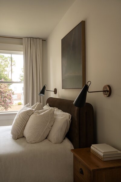 Close-up of master bed in colonial-style home with thick brown headboard, white textured bedding, gold and black sconces, neutral curtains, and moody painting above.