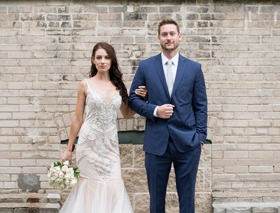 bride and groom standing against brick wall look seriously toward camera