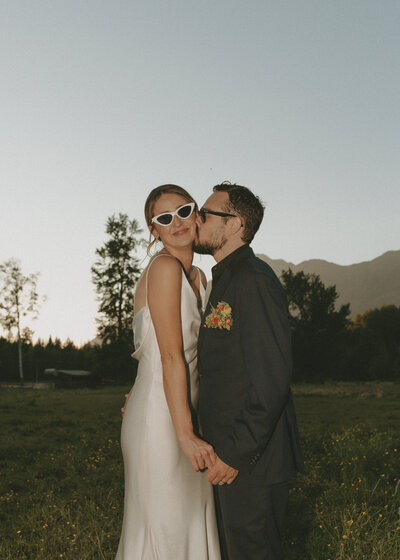 groom kissing bride on the cheek while she is smiling at the camera in the mountains