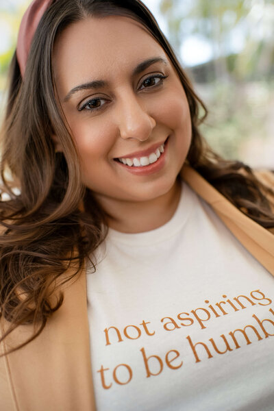 Founder Lena with long hair, wearing a headband, smiles warmly. Her shirt reads "not aspiring to be humble," conveying confidence and positivity.