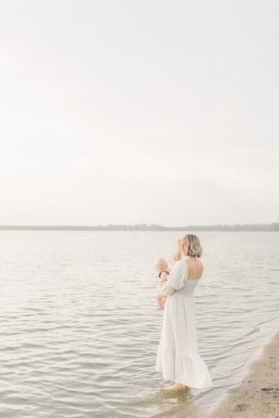 Mother standing barefoot in the lake holding her 6 month old close during a serene milestone photography session in Kansas City.