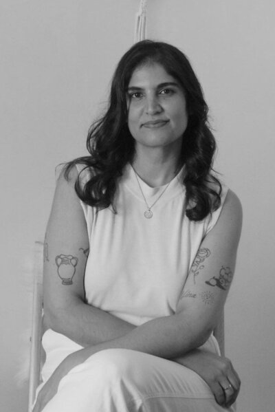 Black and white photo of woman with curly brown hair smiling at the camera wearing a white sleeveless shirt showing several tattoos on her arms.