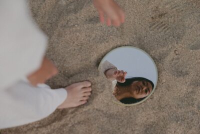 Woman experiencing mid-life crisis and betrayal, reflecting on herself through a circular mirror while standing barefoot on the sand. This image symbolizes personal reflection and freedom in the journey towards healing and transformation.