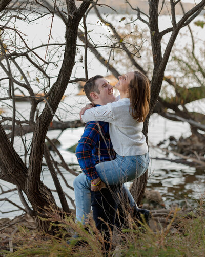 A man holds a woman and swings her around as she laughs during their engagement session. They are in front of Percy Priest Lake at Long Hunter State Park in Nashville, Tennessee.