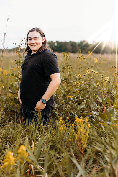 high school senior boy standing in a field with a black polo on at sunrise