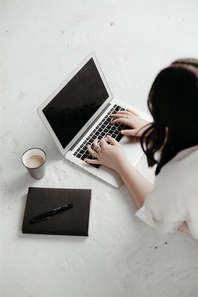 An overhead view of a professional workspace featuring a woman typing on a laptop, alongside a coffee cup and notebook, representing the meticulous and organised process offered by KP Virtual Assistance