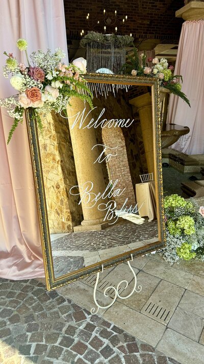 Gold vintage mirror displaying "Welcome" sign with calligraphy and flowers