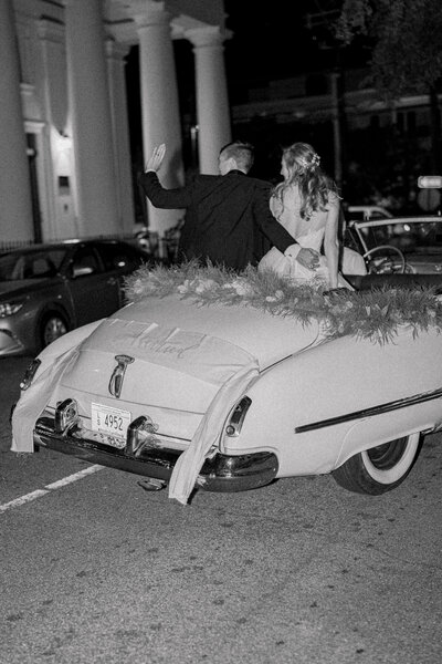 black and white photo of bride and groom in convertible getaway car. Groom waves to the crowd as car pulls away from outdoor intimate wedding in Charleston.