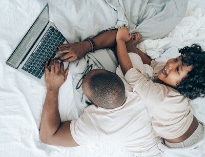 Overhead photograph of a black man and young boy on a bed with a laptop. Photo by Ketut Subiyanto via Pexels.