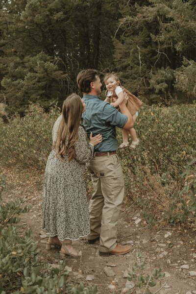 Photo taken by Bailee Freeman Photography of mom, dad, and baby girl in a forest of green trees with yellow flowers standing on a dirt path. Dad is lifting baby girl into the air to get a smile and mom is admiring them. 