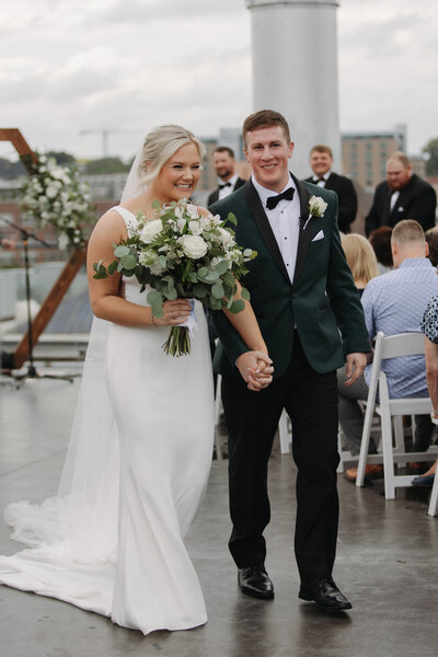 The bride playfully runs away as the groom chases her. She is dressed in a mid length satin white dress and has a short veil flowing behind her.