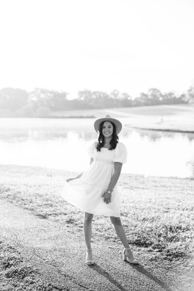 Black and White image of senior in white dress with hat in front of lake