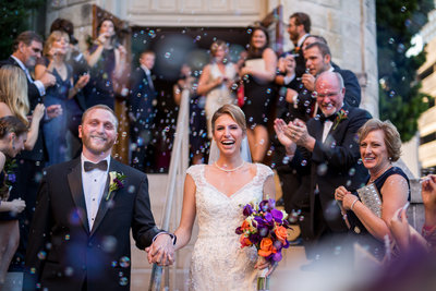 Bride and groom with big smiles exiting a church surrounded by bubbles.
