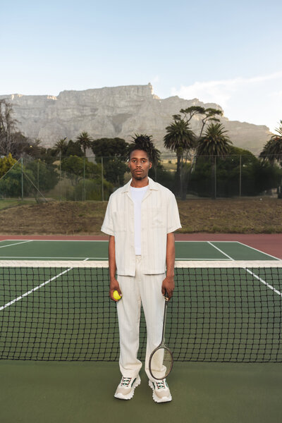 A stylish man in a cream outfit holding a tennis racket and ball, standing confidently on a tennis court in Cape Town with Table Mountain and palm trees in the background.