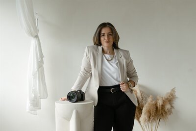 Documentary and editorial wedding and elopement photographer in Orlando, Florida, posing in a minimalist studio with a Canon camera, wearing a beige blazer, white shirt, and black pants.
