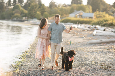 Engaged couple with dog walking on Bainbridge Island waterfront at sunset.