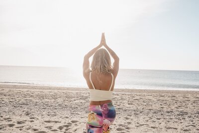 Yoga teacher on the beach, seen from behind, with hands raised above her head in a prayer pose, leading a group class.