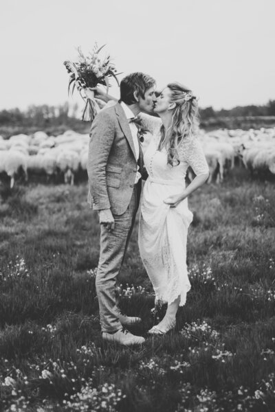 Bruidspaar kust elkaar met het bruidsboeket in de lucht. Ze staan voor een schaapskudde op de heide in Elspeet op de Veluwe. Ze tilt haar vintage jurk wat omhoog en je ziet haar trouwschoenen. De bruidegom draagt een pak met een strik.