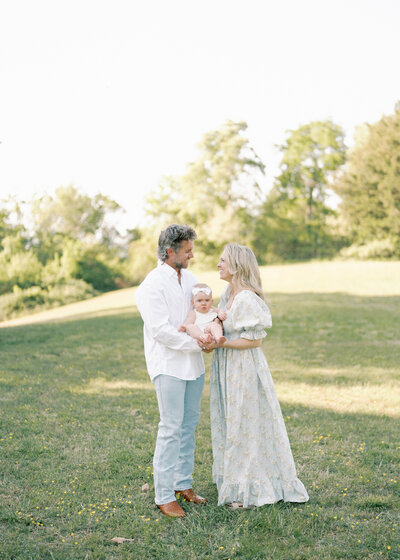 Woman wearing black floral dress and man wearing white button up shirt hold their daughter on a beach with trees in the background.