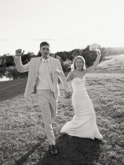 Bride and groom joyfully walking hand in hand through an open field after their wedding ceremony.