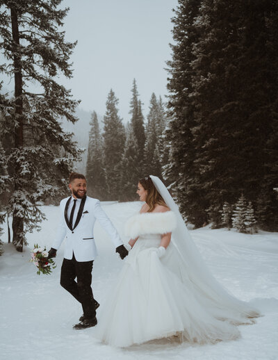 the bride and groom are walking in the snow together with tall pine trees behind them. they are smiling and laughing.