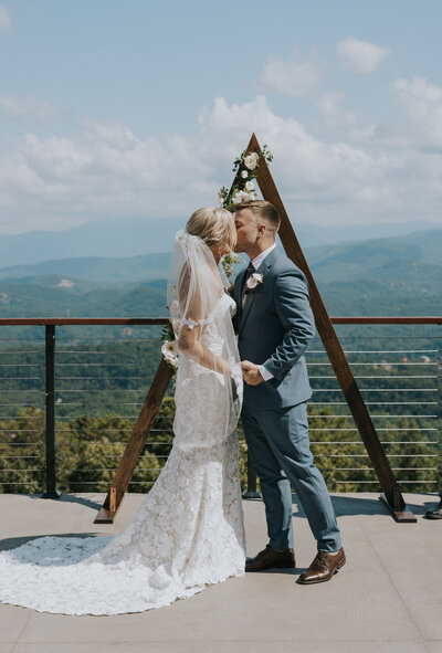 smoky mountain wedding with bride and groom holding hands and standing at a triangle arch on a deck with the smoky mountains in the distance as they kiss at the end of their ceremony photographed by Photographer Knoxville TN