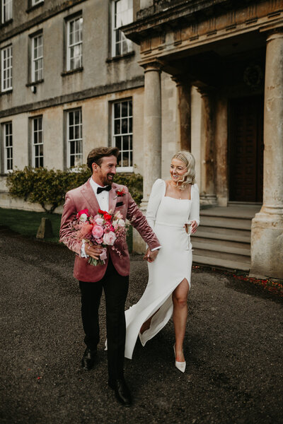 A groom carries the bouquet and holds his brides hand as they walk in front of the main house at Elmore Court. 