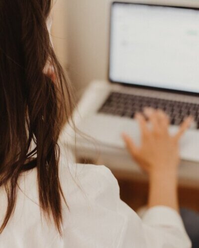 Woman working on a laptop at a desk, seen from behind with long brown hair and a white blouse.