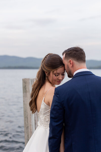 Bride and groom rest their heads against each other on the dock of a lake