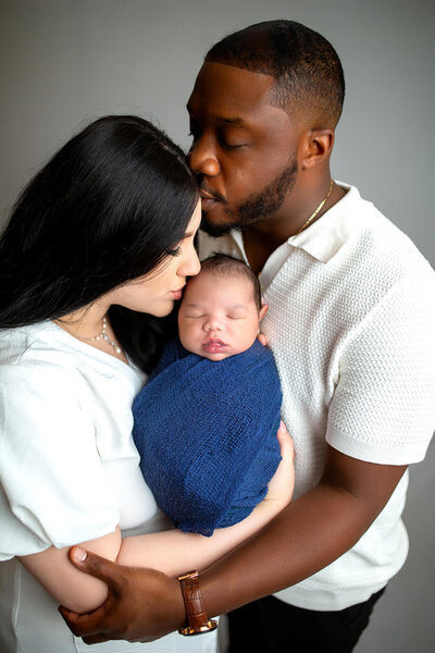 Studio newborn session with mother holding baby boy and kissing side of baby's head with father's arms wrapped around mother and baby kissing mothers forehead 