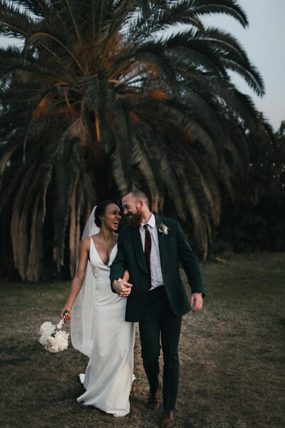 Bride holding her white floral bouquet while walking and holding her groom's hand and laughing