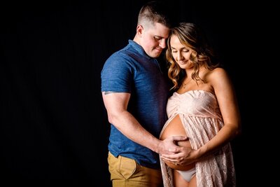 A tender maternity portrait of an expectant couple: the man embraces the woman from behind, both looking down at her pregnant belly with hands gently placed, against a classic dark backdrop