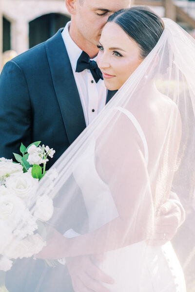 Close up image of groom kissing bride on the head while her veil blows in the wind
