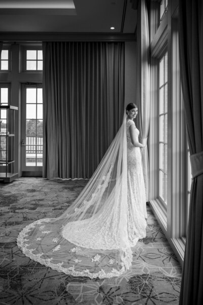 Black and white bridal portrait taken indoors at Carmel Country Club in Charlotte, NC. The bride is partially facing away, looking back over her shoulder toward the camera, smiling. Her cathedral veil with lace detailing is beautifully laid out behind her.