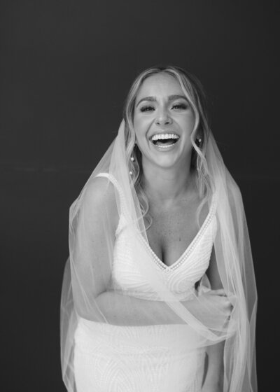 Black and white close up portrait of bride smiling and holding her veil in front of a dark studio background
